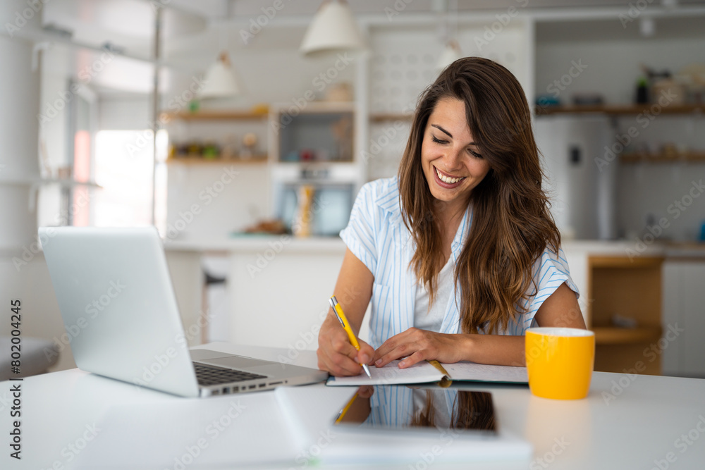 Smiling young woman using laptop sitting at the table in a home office. Focused female entrepreneur in casual clothing looking at the paper, writing down ideas or a future business plan. Smiling young woman using laptop sitting at the table in a home office. Focused female entrepreneur in casual clothing looking at the paper, writing down ideas or a future business plan.