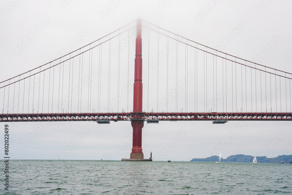 Naklejka premium Iconic Golden Gate Bridge view from water level in San Francisco, California. International Orange color pops against overcast sky, boats in distance. Engineering marvel captured.