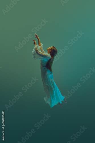 Woman dressed in flowing layers of white extends her arms towards sky from water depth. Model levitating against aqua background. Concept of serenity and calm, freedom, dream, surreality, fantasy. Ad