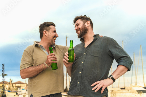 Two men - laughing and holding beers at marina - casual atmosphere, friendship moment, harbor background - leisure, bonding. (ID: 802024405)