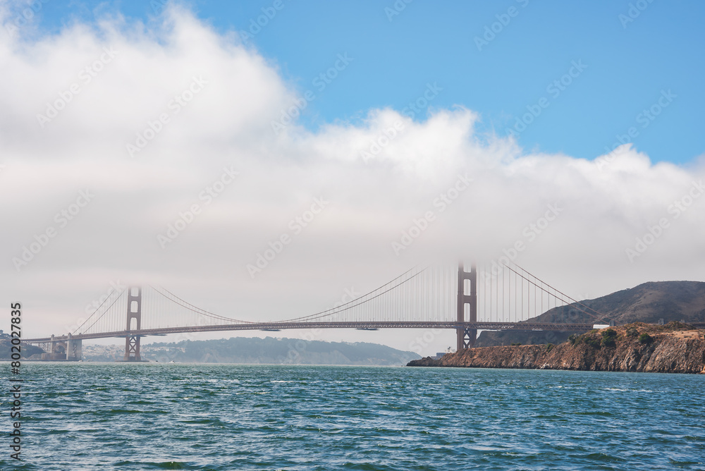 Daytime view of the Golden Gate Bridge in San Francisco, CA. Iconic orange bridge against blue sky, serene water, suspension towers, Marin Headlands frame landscape. Tranquil and majestic scene.