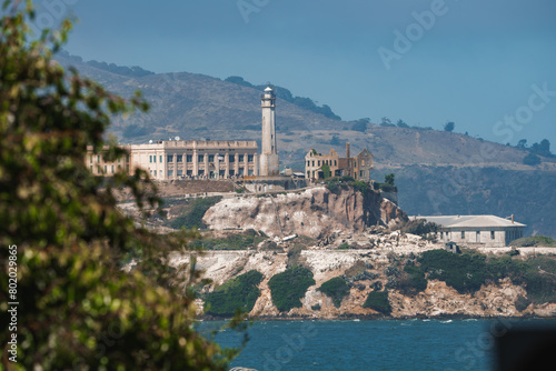 Distant view of Alcatraz Island in San Francisco Bay. Former federal prison with imposing structure and lighthouse. Rugged terrain, sparse vegetation, bay waters, clear skies.