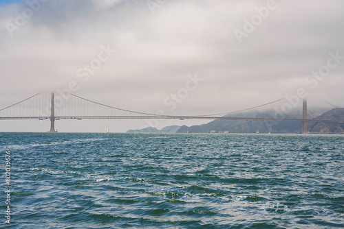 Iconic Golden Gate Bridge in San Francisco, California. Serene view from water's edge highlights vivid bridge color against overcast sky, hills in background.