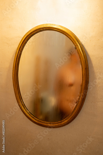 a nude woman and a wooden structure with a towel in the reflection of a fogged mirror with a golden frame, cream-colored wall of fine cement. Warm light coming from above the mirror