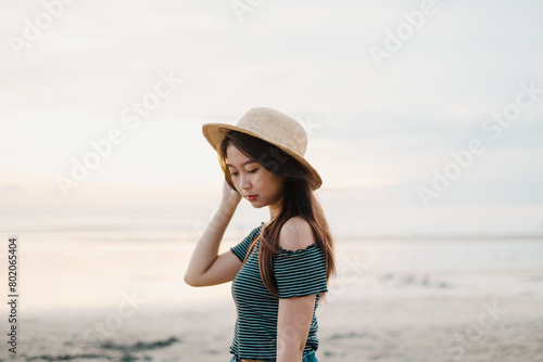 A beautiful young Asian girl is holding her straw hat on the beach during sunset with bokeh background