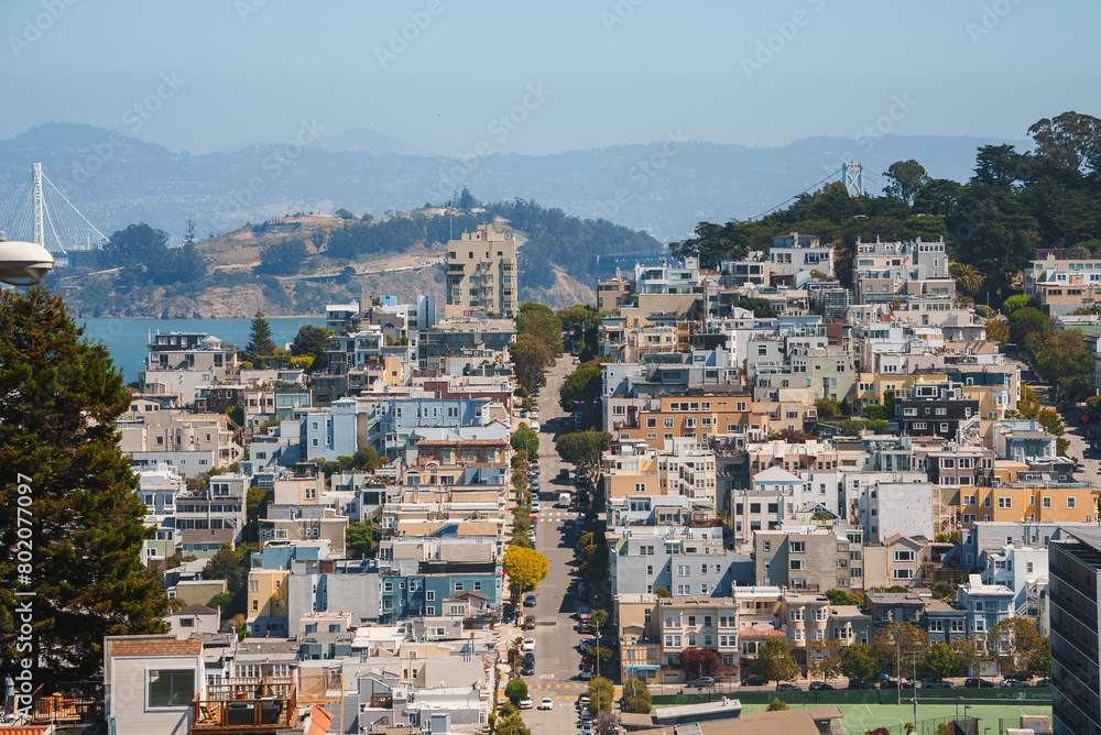 Residential area in San Francisco with bay windows, fire escapes, and hilly streets. Green hills in background, clear blue sky. Bay Bridge partially visible.