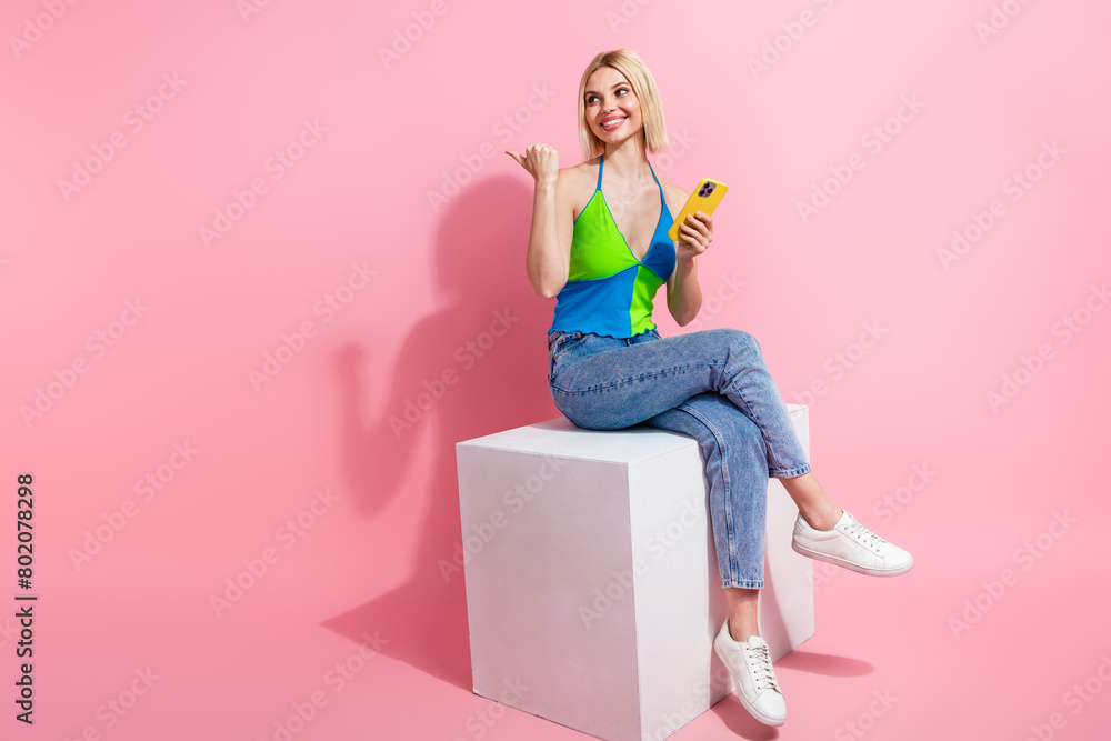 Full size photo of charming woman wear top sit on platform look directing empty space hold smartphone isolated on pink color background Full size photo of charming woman wear top sit on platform look directing empty space hold smartphone isolated on pink color background