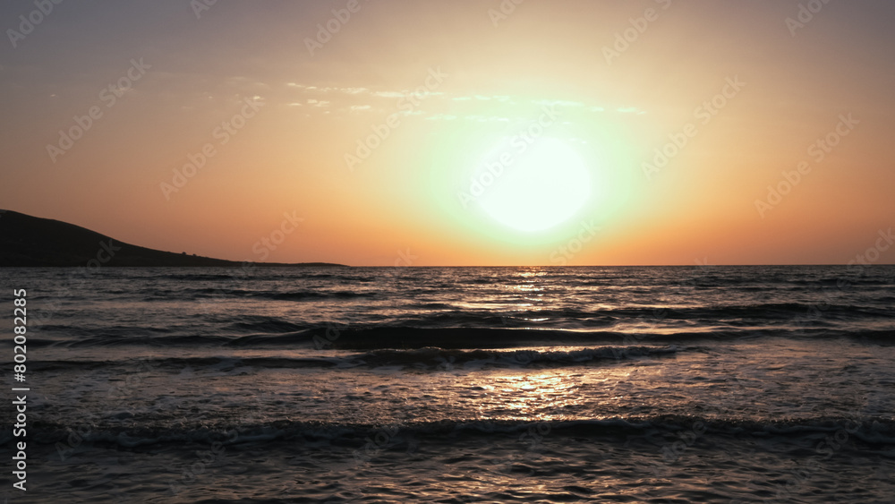The sun is setting over the ocean, casting a warm glow on the water. The sky is a mix of orange and pink hues, creating a serene and peaceful atmosphere. The calm waters reflect the colors of the sky