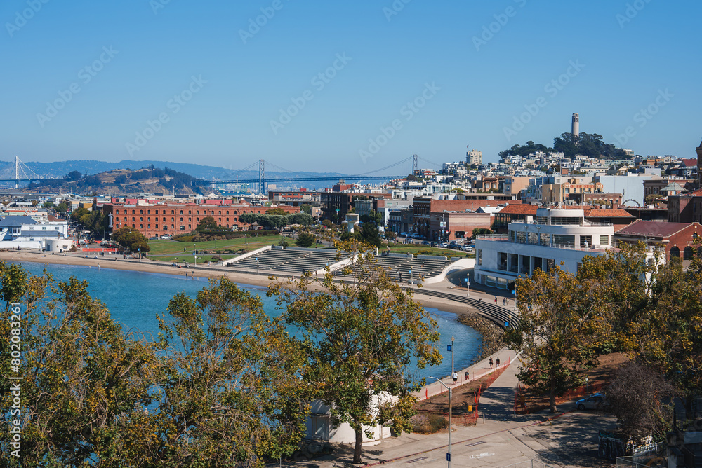 Scenic view of San Francisco's waterfront with a park, buildings, Coit Tower, Bay Bridge, and clear blue sky. Vibrant urban area for leisure and recreation.