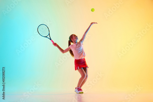 Teenage athlete girl, tennis player serving ball in motion perfectly to start tennis match in neon light against blue-yellow background. Concept of individual kind of sports, fashion, tournament. Ad © Lustre