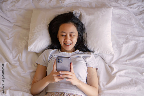 Top view of happy asian young woman awaken in white cozy bed using modern smartphone gadget, happy millennial female wake up lie in comfortable bedroom browsing Internet on cellphone in the morning