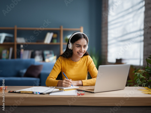 Happy Indian girl in wireless headphones watching learning webinar, online lecture on laptop, speaking at educational conference, writing notes at table, studying on Internet from home