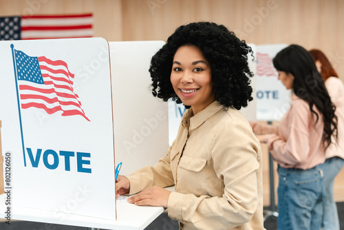 Smiling beautiful African American woman with curly hair standing in booth with American flag photo