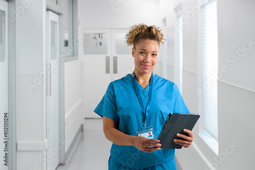 Portrait Of Smiling Female Nurse Wearing Scrubs In Hospital Corridor With Digital Tablet