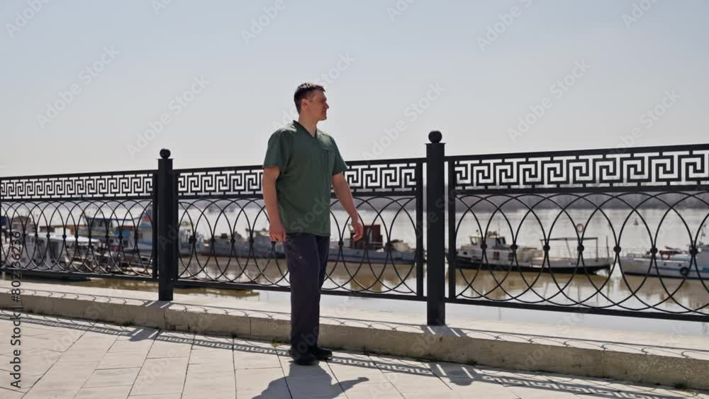 The doctor walks along the promenade in sunny weather. A man walks at a leisurely pace in slow motion. Confident man in green scrubs walking along a riverside fence with urban skyline and river in the The doctor walks along the promenade in sunny weather. A man walks at a leisurely pace in slow motion. Confident man in green scrubs walking along a riverside fence with urban skyline and river in the