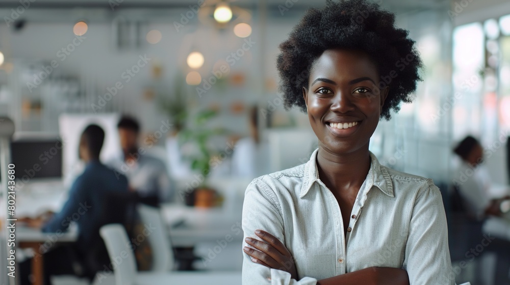 Fototapeta premium Happy Small Business Owner Black African Woman Posing with Hands Folded - Employees Working in Modern Office Behind. Millennial Female Team Leader at Workplace