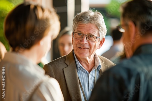 A middleaged businessman wearing glasses engaging in conversation with a group of people at a networking event, showcasing interpersonal skills and relationship-building photo