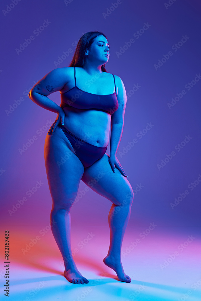 Portrayal of body positivity. Beautiful young girl with plus size body shape, posing underwear against purple background in neon light. Concept of natural beauty, body positivity, care, acceptance