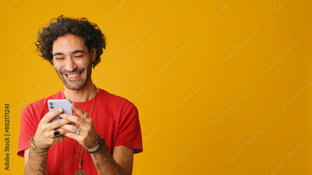 Smiling man with curly hair, dressed in red T-shirt, using mobile phone, typing message isolated on yellow background in studio Smiling man with curly hair, dressed in red T-shirt, using mobile phone, typing message isolated on yellow background in studio