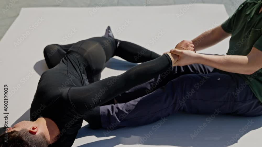 Man in green shirt performing arm stretch on person lying on mat outdoors during physical therapy session. Man in green shirt performing arm stretch on person lying on mat outdoors during physical therapy session.