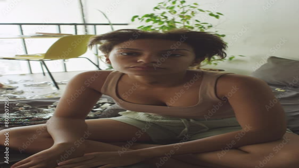 Vertical portrait shot of drowsy teen girl sitting on unmade bed in cluttered room, rubbing her face and looking at camera Vertical portrait shot of drowsy teen girl sitting on unmade bed in cluttered room, rubbing her face and looking at camera