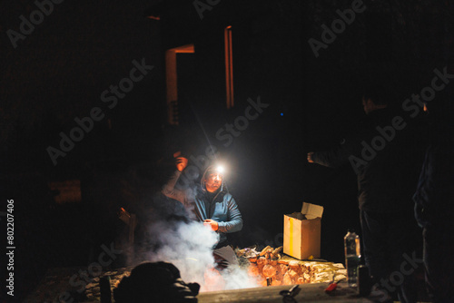 A diverse group of people stand circled around a roaring fire, their silhouettes flickering in the warm glow