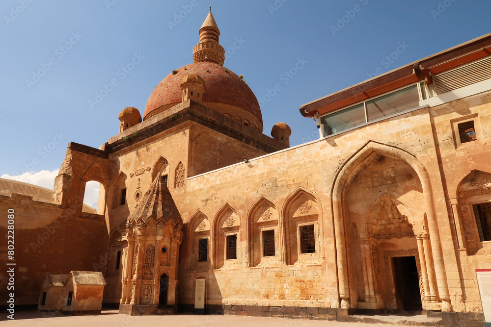Obraz premium Ottoman tomb next to a row of barred windows and a gate with the mosque with its red cupola, dome in the background, in the second court inside the Ishak Pasha Palace, Sarayi, Dogubeyazit, Turkey