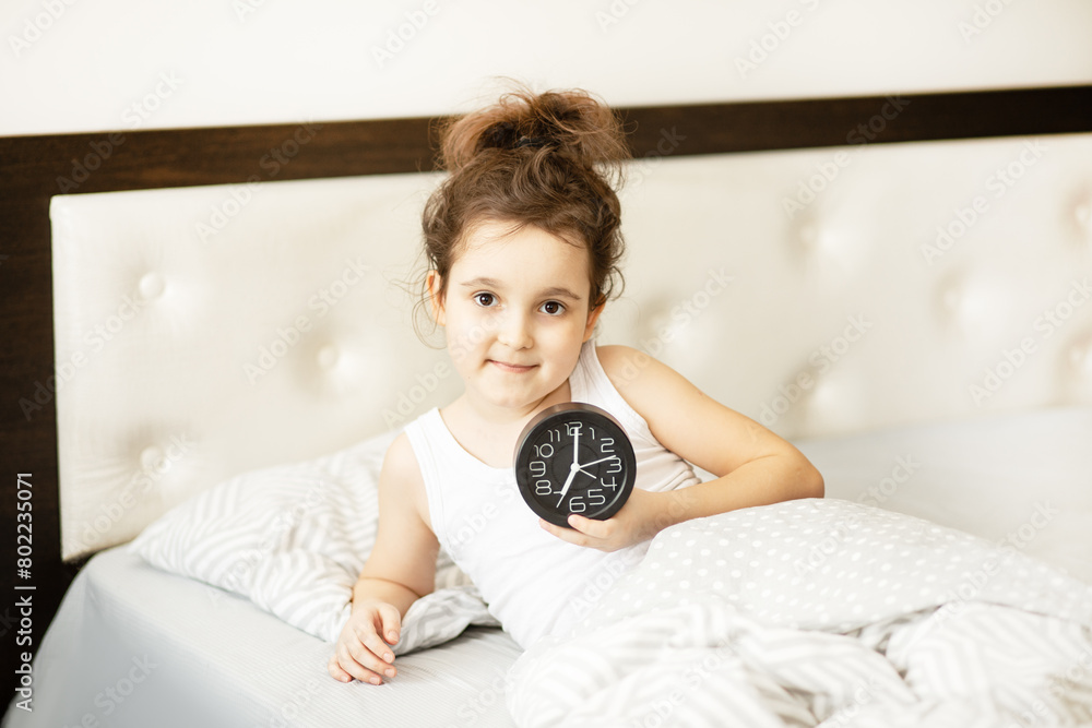 Cute little kid girl lying on bed waking up in morning and holding alarm clock in a hand. Cute little kid girl lying on bed waking up in morning and holding alarm clock in a hand.