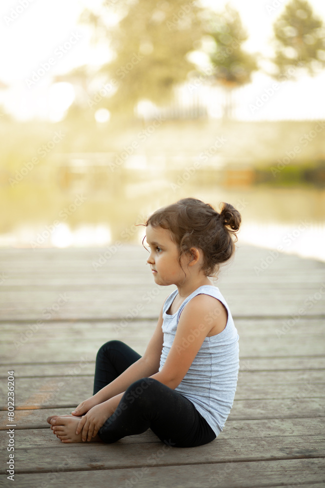 Little girl sitting in nature near by river lake and practicing yoga or resting after sports exercises. Healthy lifestyle. Little girl sitting in nature near by river lake and practicing yoga or resting after sports exercises. Healthy lifestyle.