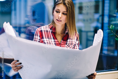 Professional young architect concentrated on reading blueprint of project construction working overtime in office,skilled female student holding big paper with graphic drawing prepare hometask photo