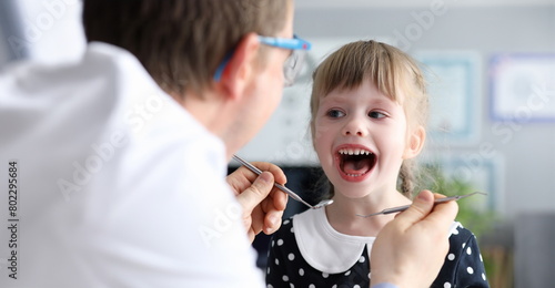 Male dentist look at open mouth litle happy girl child againlt hospital office background portrait. Tooth treatment concept