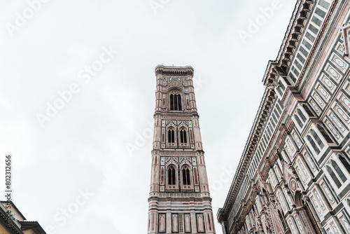 Santa Maria del Fiore's Gothic-Renaissance intricate façade framing Giotto's Campanile from Piazza del Duomo, standing tall in the centre, against the cloudy sky.