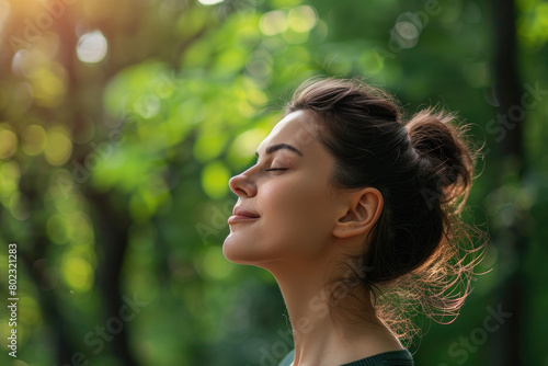 A woman breathes in fresh air with her eyes closed and looks up at the green trees behind her  enjoying nature s tranquility and feeling of well-being