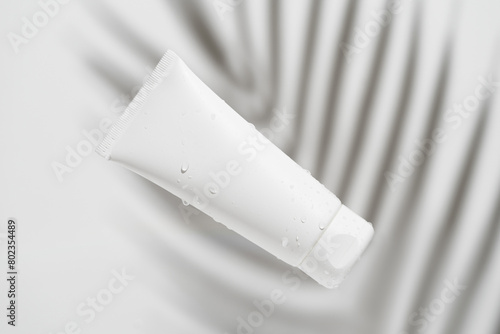 White facial cream tube mockup with water drops on palm branch shadow background on gray isolated background. The concept of moisturizing and nourishing the skin.