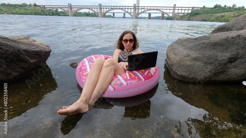 A young woman in a swimsuit works on a laptop while swimming in an inflatable circle on the river. Freelance. Remote work.