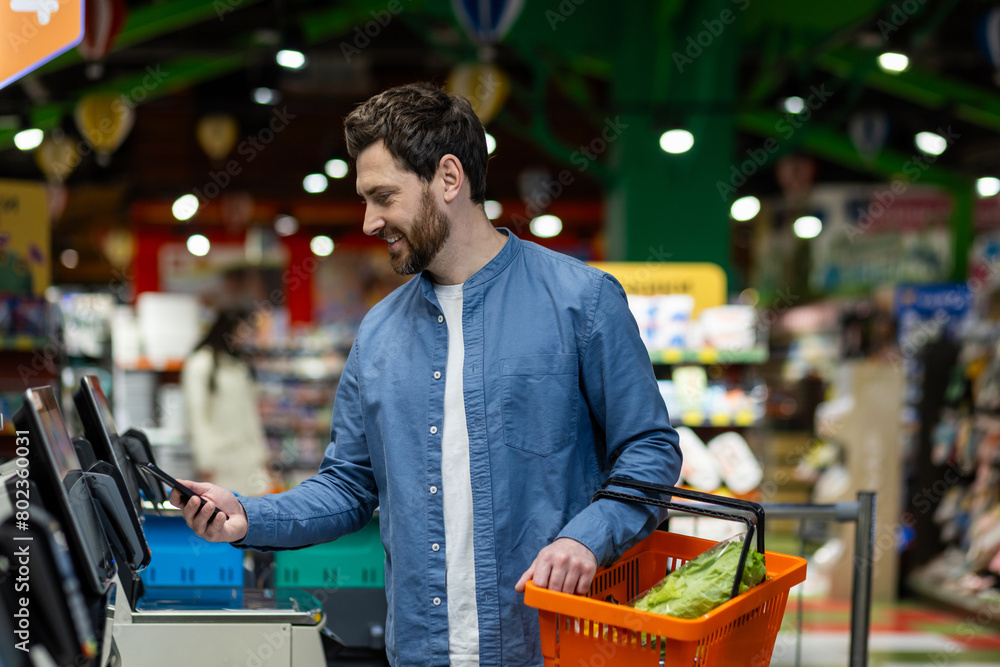 © Liubomir - Satisfied customer of food store tapping modern gadget to electronic device while carrying orange basket. Relaxed man in denim shirt enjoying benefits of using banking application for remote payment.