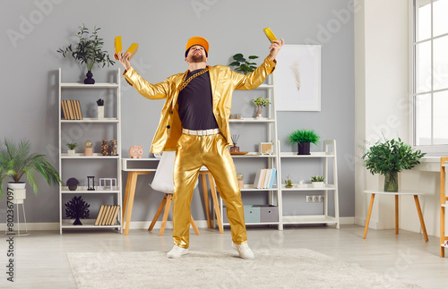 Rich man in a golden business suit celebrating financial triumph or win at home. He is holding gold bars, symbolizing wealth and success. Victorious and prosperous individual, rapper or winner. © Studio Romantic