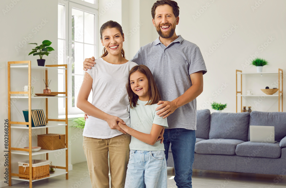 Portrait of happy loving nuclear family at home. Mother, father and child spend time together. Beautiful mom, bearded dad and pretty daughter standing in living room, looking at camera and smiling Portrait of happy loving nuclear family at home. Mother, father and child spend time together. Beautiful mom, bearded dad and pretty daughter standing in living room, looking at camera and smiling