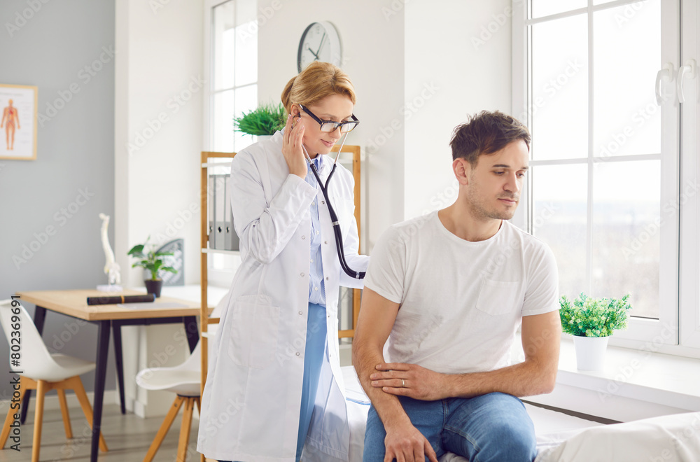 Obraz premium Medical service. Healthcare professional conducting physical examination on male patient in modern clinic setting. Focused female doctor in white coat uses stethoscope to listen to heartbeat of man.