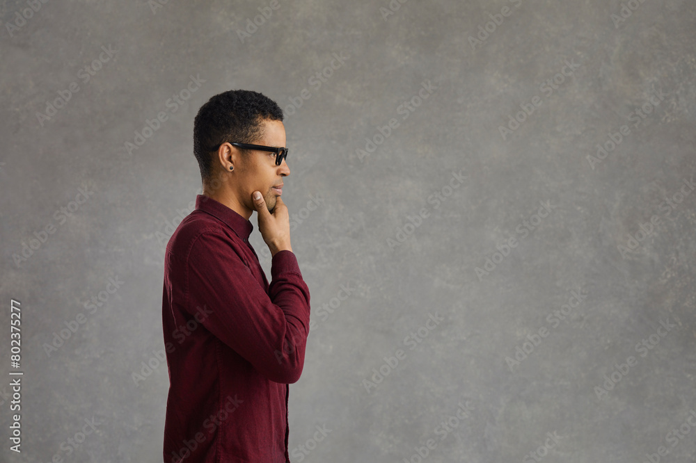 Pensive african american customer thinking over special offer, touching chin looking forward on grey studio copy space. Handsome young man in casual shirt and glasses side view portrait Pensive african american customer thinking over special offer, touching chin looking forward on grey studio copy space. Handsome young man in casual shirt and glasses side view portrait