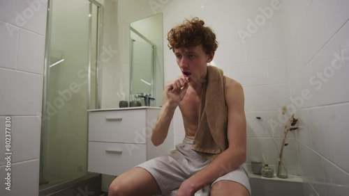 Young hungover man sitting on toilet in bathroom, brushing teeth and wiping mouth with towel while feeling sick next morning after party