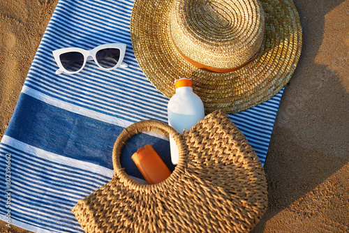 Sunbathing accessories on sandy beach, straw hat beside sunglasses, sunblock lotion in bag for skin protection, summer seaside holiday essentials on striped towel, outdoor UV care on sunny day.