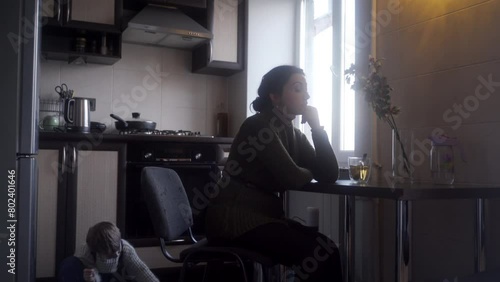 Worried mother sits at kitchen table, contemplating, as child plays quietly behind her. Domestic life with subtle tension, family issues, indoor, natural light.