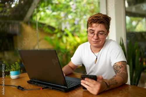 Transgender office worker with tattoos checks smartphone, distracted from laptop. Casual business setting, gender diversity at work. Indoor plant, natural light, inclusion in modern workplace.