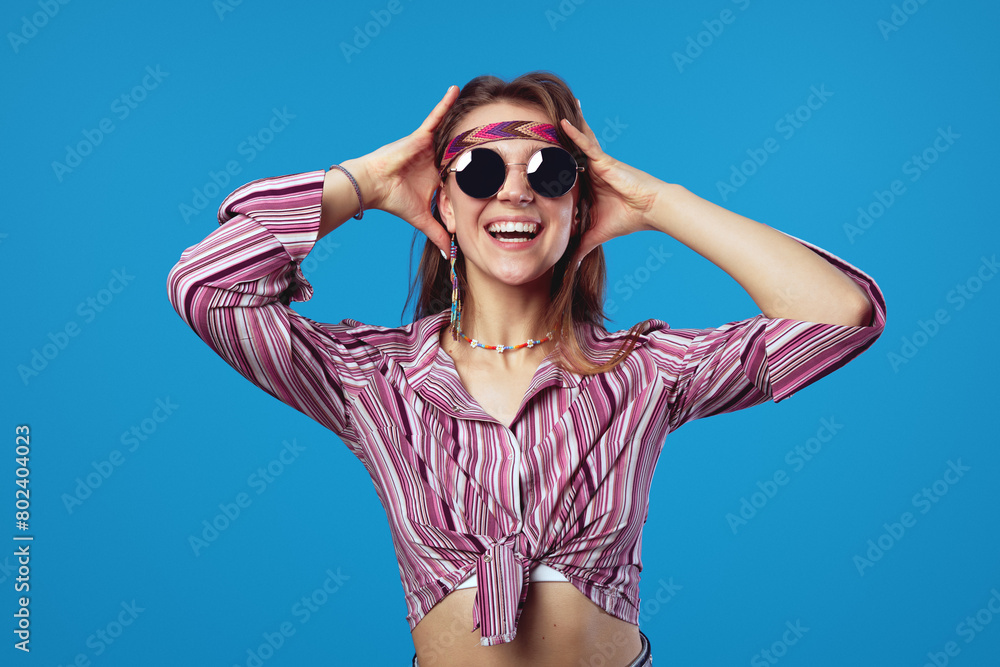 Joyful happy excited hippie young woman in trendy sunglasses, holding hands on head and smiles broadly, isolated blue background