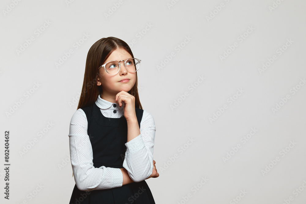 Clever primary kid girl with glasses holding hand under the chin and looking away at free space while thinking, isolated over white background Clever primary kid girl with glasses holding hand under the chin and looking away at free space while thinking, isolated over white background