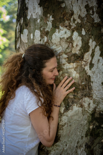 Beautiful woman with gorgeous curly hair touching the tree in nature, dressed in white,  concept: active healthy life, in love with nature, support, balance, wellbeing, energy, forest, healing 