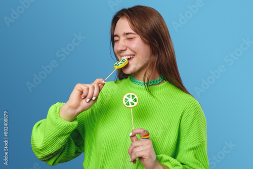 Lovely young female model keeping eyes closed and enjoying a delicious lemon lollipop while standing isolated on blue background.