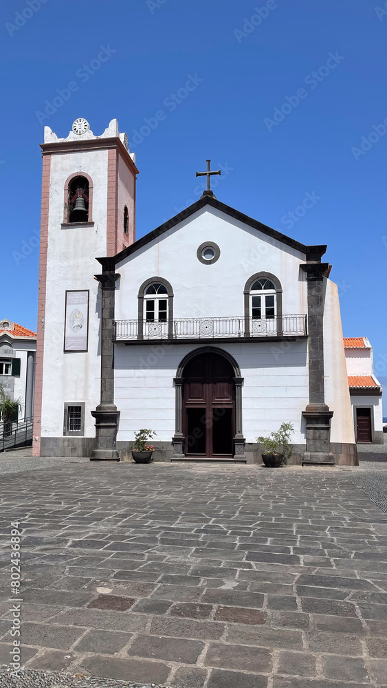 Fototapeta premium A charming white church with a bell tower in a small square on Madeira Island, standing under a clear blue sky.