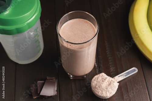 Protein milkshake cocktail in a glass, plastic measuring spoon with whey protein powder, shaker for prepare blended protein drink, chocolate cubes and banana fruit on a dark wooden background